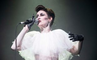 Maude Davey in 'Performing the Monster'. Davey will be delivering a master class as part of the Festival of Australian Queer Theatre. A pale-skinned woman on stage holding a microphone up to her lips. She is wearing smokey eyeshadow and black gloves with a white frilly gown.