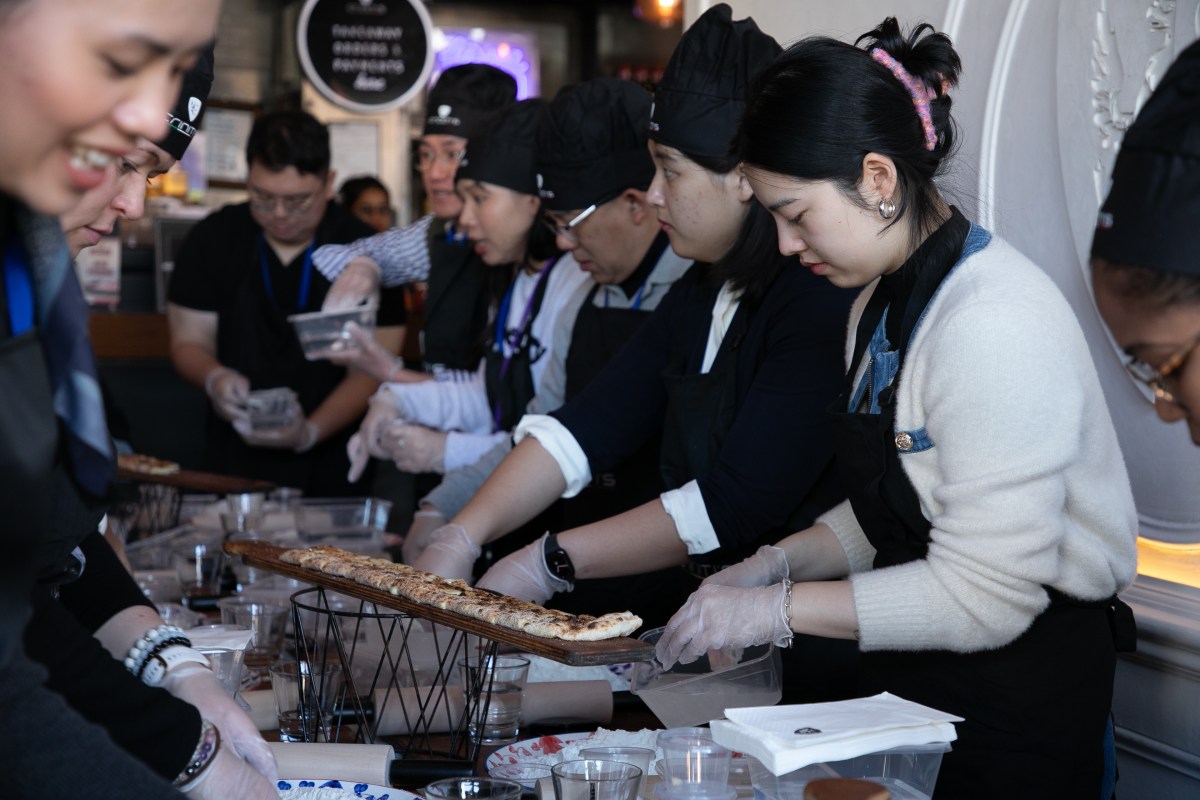 Leadership is like pizza making – the ingredients are simple but it takes time and technique to nail. A group of people lined up across two sides of a long table making pizza dough. 