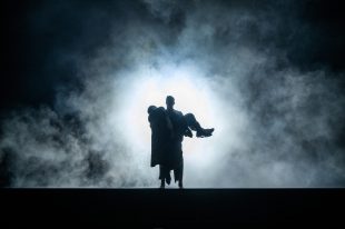 In the middle of a swirling foggy dark stage with a bright spotlight behind two silhouetted figures stand. The monster creation is carrying Dr Frankenstein in his arms.