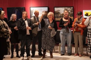 'While the World Waits' an Arts Outwest curated exhibition recently opened at NSW Parliament House. A group of people standing in front of several artworks hung on a burgundy coloured wall. They are clapping and looking towards a middle-aged woman standing in the centre waving her hand at the camera.