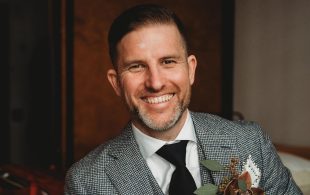 Sam Clark has died aged 40. Image is a Caucasian man with slicked back hair, a clipped greying beard and a big smile. He is wearing a white shirt, dark tie and a grey suit jacket.