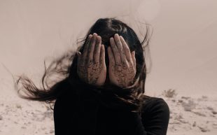 Second Generation Collective awarded $100,000 through Creative Australia’s VACS Major Commissioning Projects Fund/PICA. A woman with flow brown hair swept up by the wind. She is hinding her face behind her hands with Farsi text. She is standing in a desert.