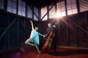 Silence & Rapture. A female dancer with slicked back dark hair has one leg bent with her foot touching her other knee and one arm stretching up and the other across to a bald headed man standing and playing a double bass. They are in a wharf building converted to a studio/stage space.