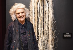 Woman with grey hair and smiling wearing black next to textile art work. Liz Williamson.