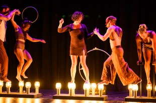 Five performers in shades of brown are walking on top of candle jars.