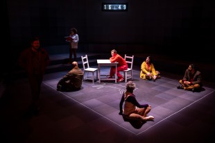 A teenage boy in red is seated a table with two chairs. Around him are four people seated on the edge of the rectangular platform while two other people are standing in the shadows.