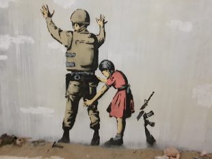 Banksy. Image is a stencil on a wall of a soldier spread against the wall with his arms up while a young girl in a pink dress frisks him and a broken rifle is next to her.