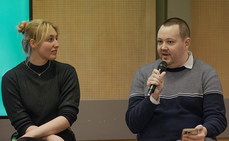 NIDA. A white woman in a black sweater with her arms crossed and a blond bun is listening to a man in a gray and black sweater who is speaking into a microphone. They are both seated.