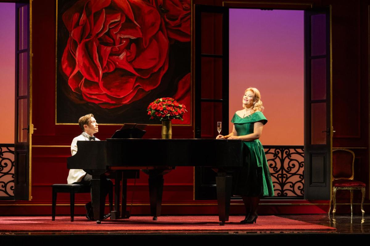 La Rondine. A man in a white tuxedo is sitting a piano. On the right is a blonde woman in a green gown next to it. There is a bouquet of roses on the piano and a large scale picture of roses in the background.