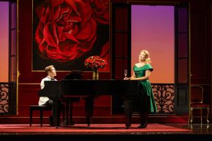 La Rondine. A man in a white tuxedo is sitting a piano. On the right is a blonde woman in a green gown next to it. There is a bouquet of roses on the piano and a large scale picture of roses in the background.