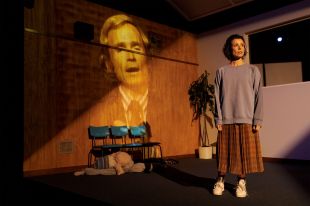 A dark-haired white woman wearing a grey jumper, a brown checked dress and white shoes stands onstage in what could be a bland office set. Three blue chairs are lined up against the rear wall, upon which is projected the face of an older man in a suit and toe. A dying pot plant stands in one corner of the room.