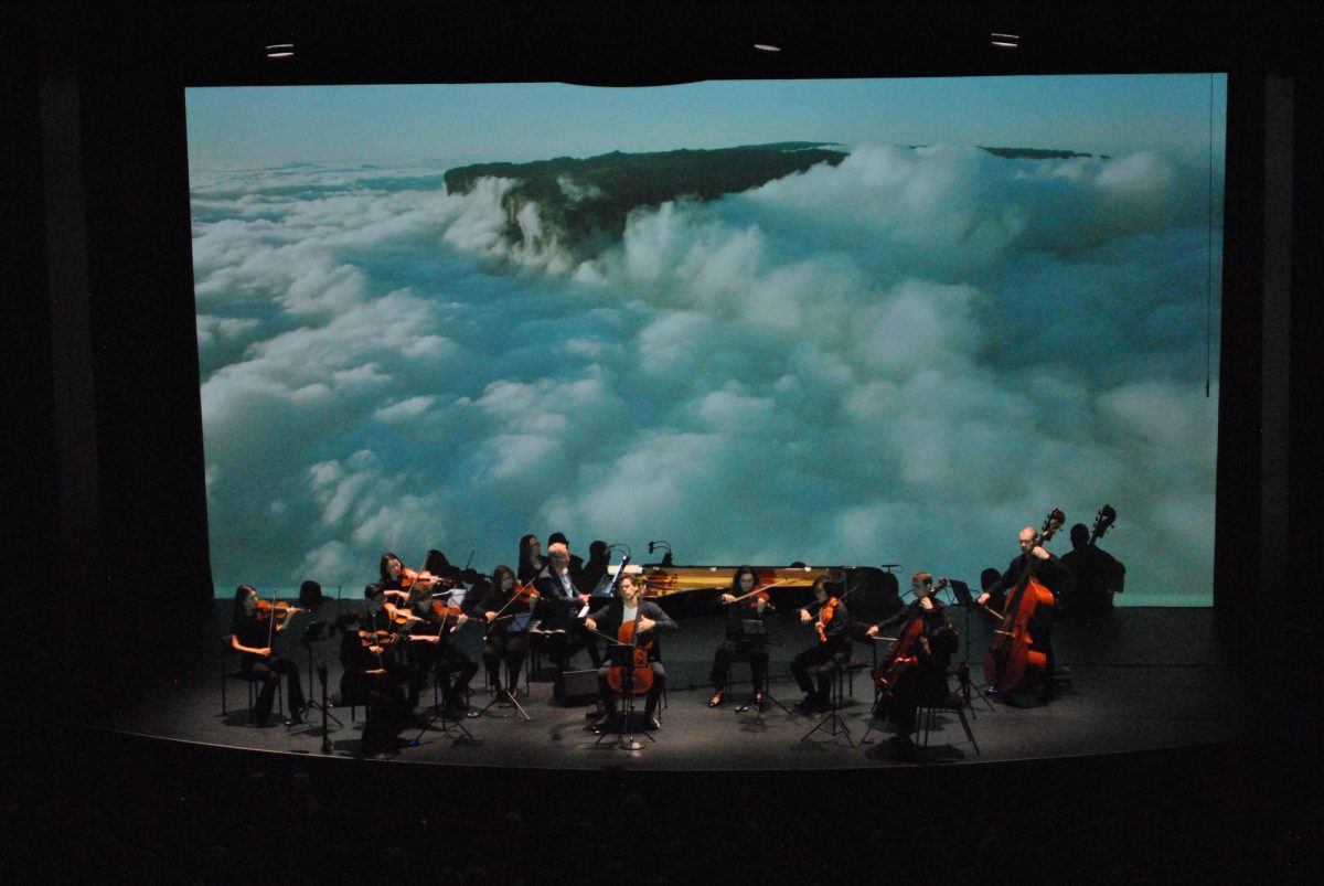 Wonderful World. An orchestra is playing on the stage with a cellist front and centre, spotlit. Behind them is a large screen of a tract of land peeking through what could be either clouds or crashing waves.