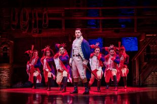 A dark-haired man in a colonial American military uniform, complete with cavalry sabre at his side, faces towards the camera. A group of dancers in similar uniforms pose dramatically behind him.