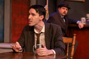 A young man in a brown suit and tie is sitting in the foreground. In the background is an older man looking at him. Between them there is a noose hanging.