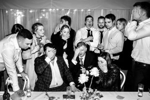 A black and white shot of a group of people at a wedding some sitting at a table, some standing and all looking on tenterhooks or stressed by something they're watching/listening to.