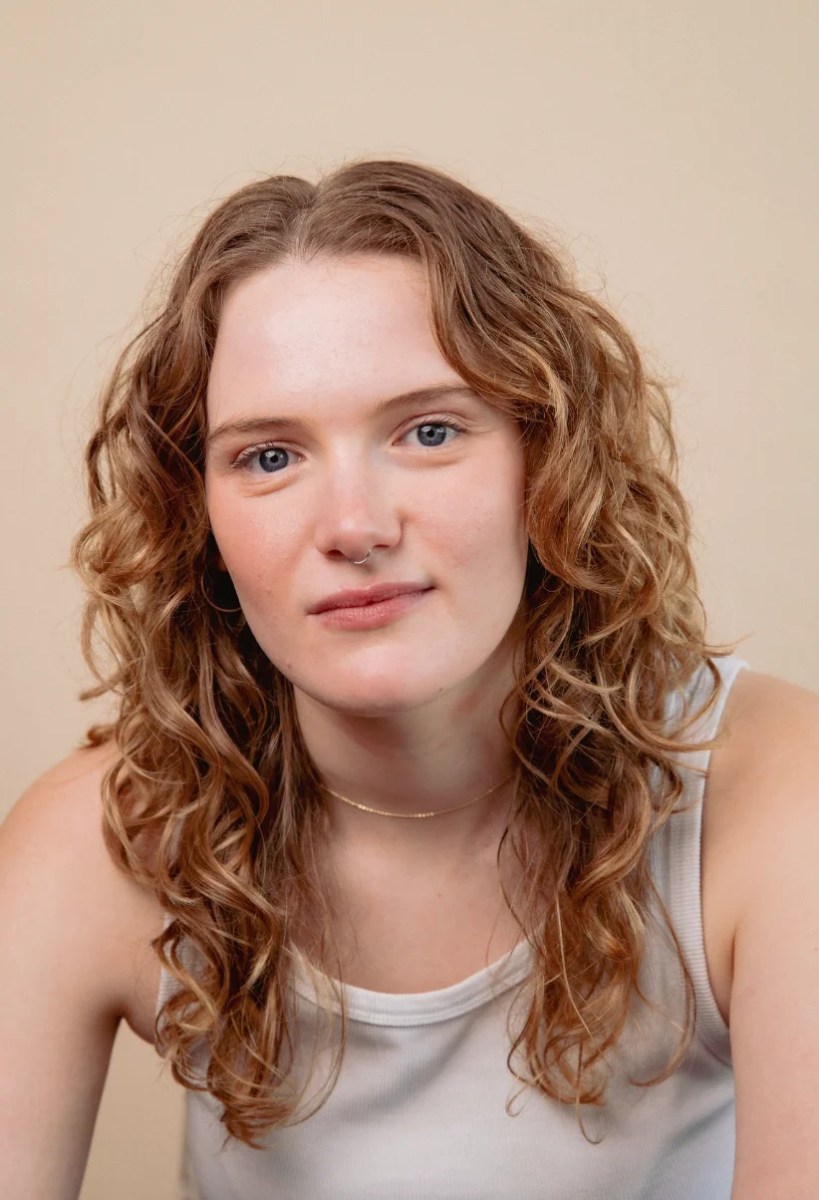 A headshot of a young Caucasian performer with long wavy fair hair and wearing a white vest. Myfanwy Hocking.