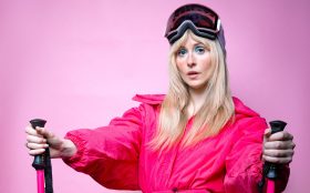 A blonde woman with ski goggles on her head wearing a pink skiing outfit.