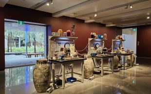 ‘Candice Lin: The Sex Life of Stone’, installation view at MUMA. An installation resembling three work stations with different lab equipment and large ceramic vessels beside each station. The space is filled with natural light thanks to a floor to ceiling window on the left side. The walls of the gallery space are paintings in a dark mahogany colour.