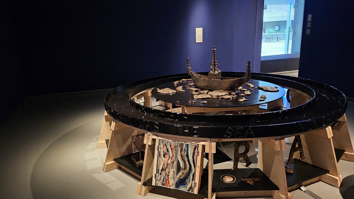 ‘I Breathe through My Anus (Night Stone)’, 2024, installation view as part of ‘Candice Lin: The Sex Life of Stone’ at MUMA. A ceramic boat glazed in oily black pigment turns slowly on a circular timber structure that channels water around its circumference. There are also letters such as ‘R’ and ‘A’ below the timbre structure. The walls of this gallery space are painted indigo. 