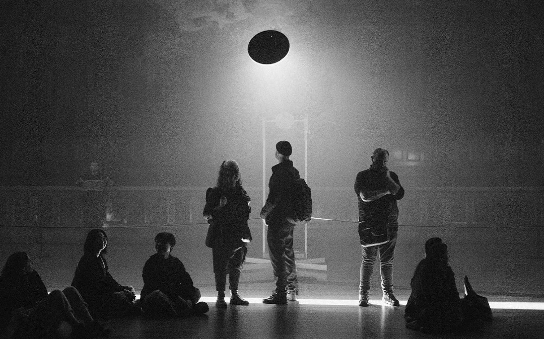 'Silent Symphony' by United Visual Artists shown in Melbourne Town Hall as part of Now or Never 2024. Image shows silhouettes of a handful of people in a large hall filled with dry ice and a mechanical installation with light coming from behind a high disc, like a UFO