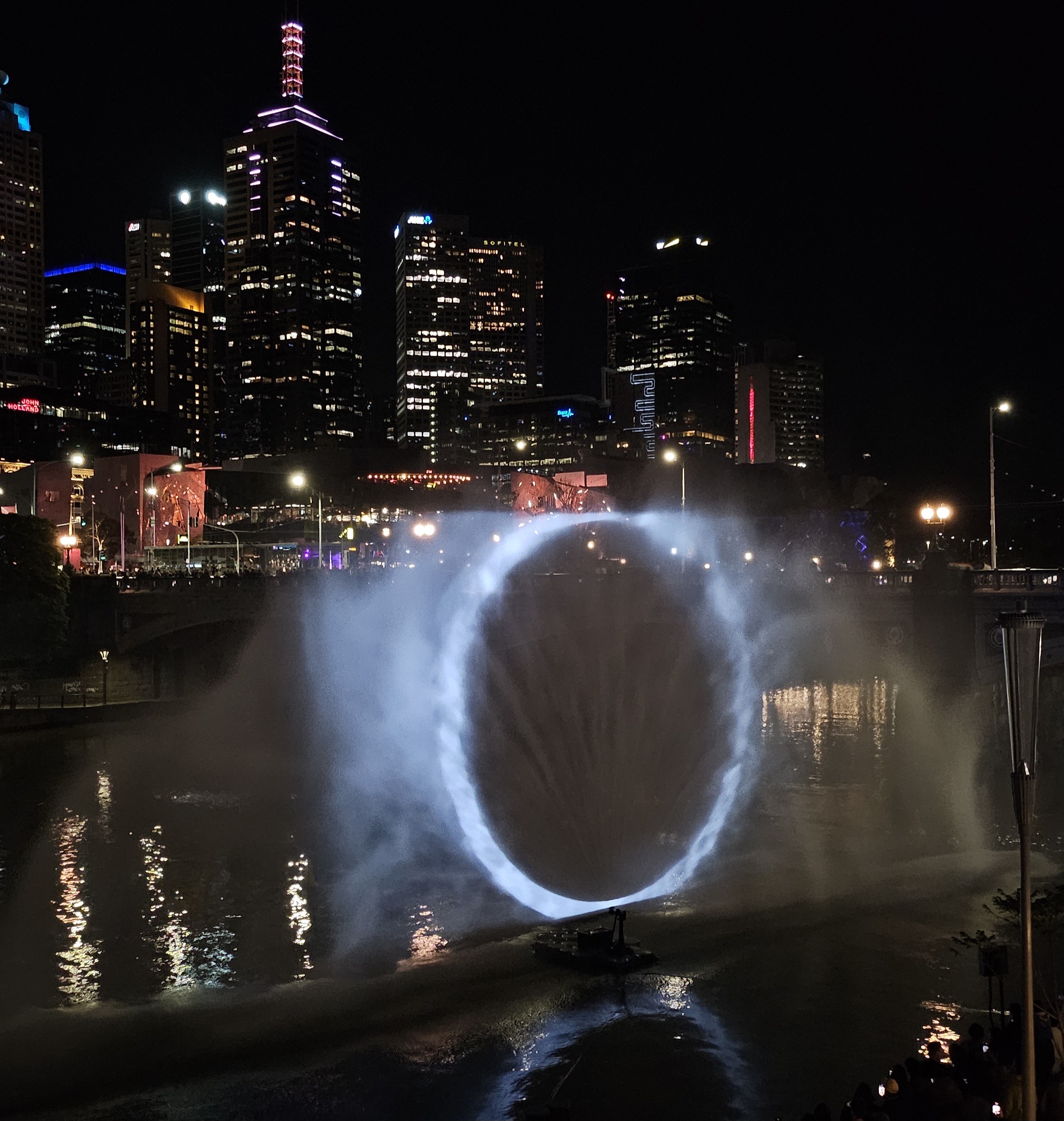 A river at night, with the city behind. Over the river is a blue circle of light projected onto water sprays, Constellations as part of Now or Never.