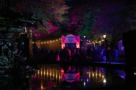 Audiences gather outside a brightly-lit venue in Prince's Square. They, the building and the night sky are reflected in a dark pool of water in the foreground.