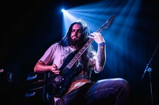 A man with long dark hair and a short dark beard concentrates as he plays the electric guitar. He wears blue jeans and a t-shirt that exposes the tattoos on his forearms.