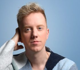 A blond man wearing a jumper in shades of blue.