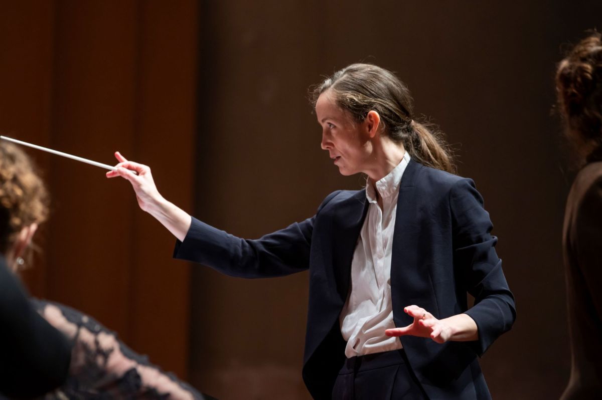 Jessica Cottis, a fair-skinned woman with long dark hair tied back behind her head, conducts the Canberra Symphony Orchestra. She wears a black jacket over a white shirt, and holds a white baton in her raised right hand.