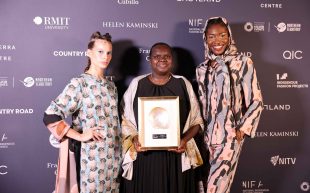 Leila Gurruwiwi accepts award for Ngali, Business Achievement Award at National Indigenous Fashion Awards 2024. Leila is a woman with dark brown skin and closely shaved black hair, wearing a black dress and earthy coloured shawl, holding the award certificate with a large shell at the centre. She is surrounded by a model on each side wearing patterened garments in front of a photo wall.