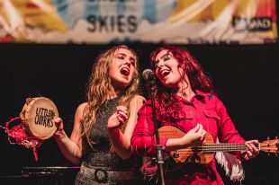 Two young female performers, one blonde-haired and playing a tambourine, the other red-haired and playing a ukulele, sing into the same microphone on stage at a music festival.