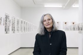 Woman with grey straight hair and black top standing in art gallery. Julie Rrap.