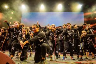 A group of young Aboriginal women dressed in black tracksuits bearing traditional designs in white, crouch and stand on stage as they sing.