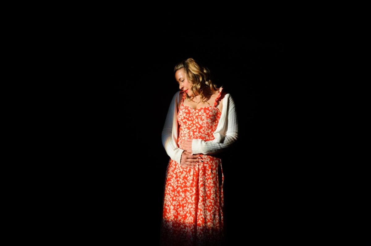 A blonde woman in a red and white print dress and a white cardigan is standing with her head bent to one side and looking down.