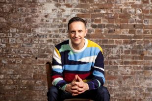 A dark-haired man wearing a vibrant, multi-coloured jumper, smiles at the camera. He sits in a wooden chair with his hands clasped, against a brick wall background.