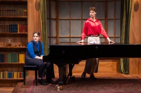 A woman wearing a blue shirt and a dark vest and pants is sitting at a piano. A man in a red shirt and light trousers is standing by the piano.