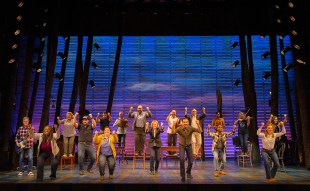 NIDA. A group of musical theatre performers are spread out on a stage with their arms stretched out. Some are standing on chairs or a table. Behind them is a dark blue backdrop.