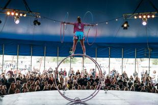 A woman in a red top and blue pleated skirt, facing away from the camera, balances atop a cyr wheel (a piece of circus apparatus) while spinning two hula hoops on each outstretched arm. In the background, a large audience watches on.