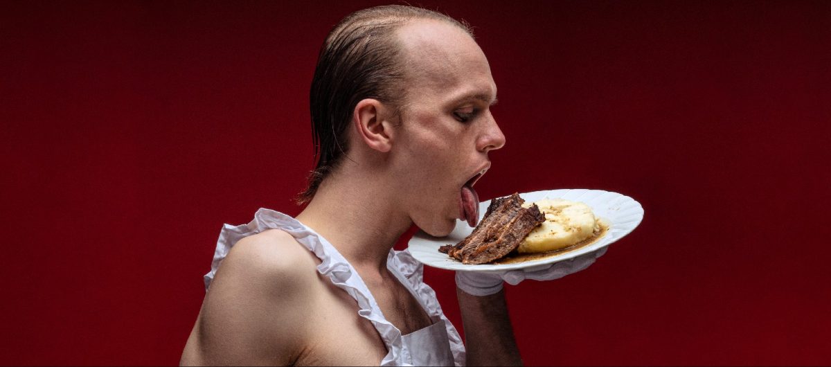 A man wearing only a frilly white apron licks a plate of chops and mashed potato, which he holds up to his mouth with one gloved hand.