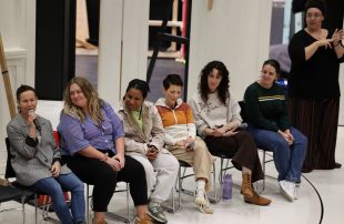 Five diverse women sit in chairs on stage as a sixth seated woman speaks into a microphone. A black-clad Auslan interpreter stands at stage left, beside one of the actors.