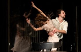 A woman in a white dress flails in the arms of a man in a white shirt. Her arms are raised, her hair flying; we cannot see her face, although she is partially reflected in pane of glass behind her.