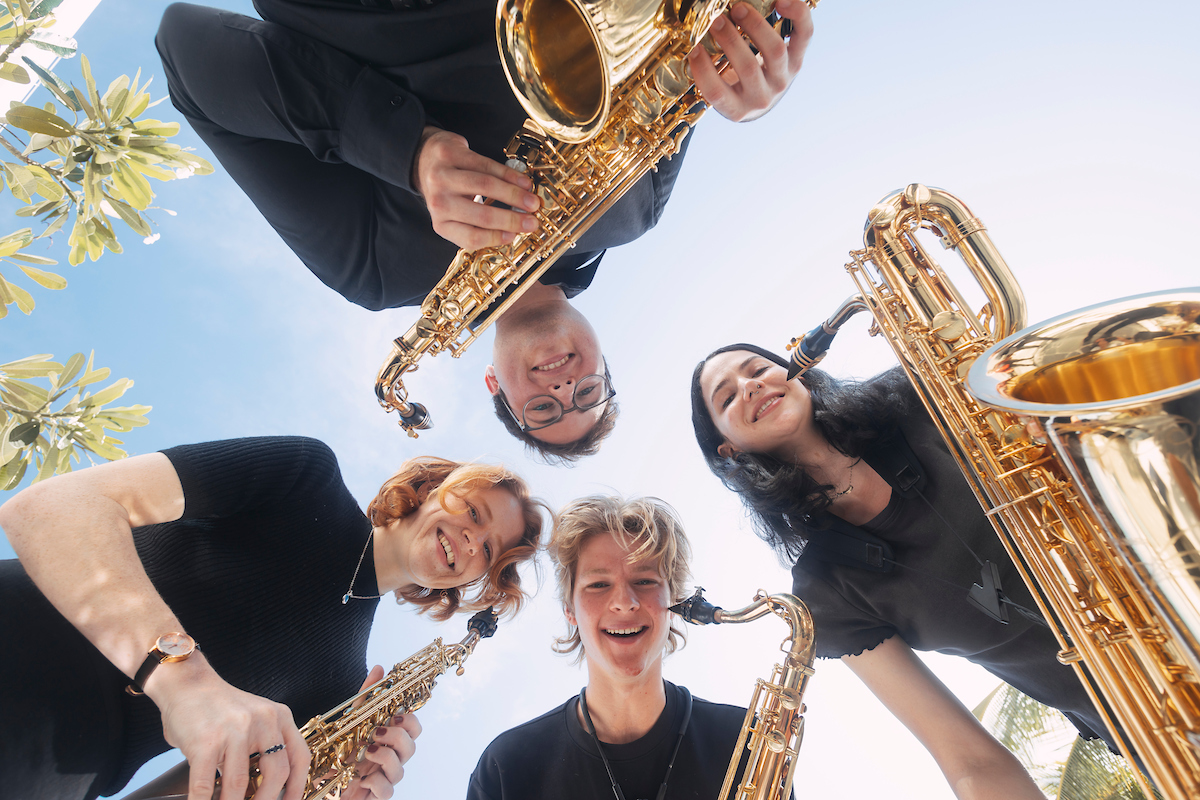 Four people in black are huddled together, looking down, each one holding onto saxophones.