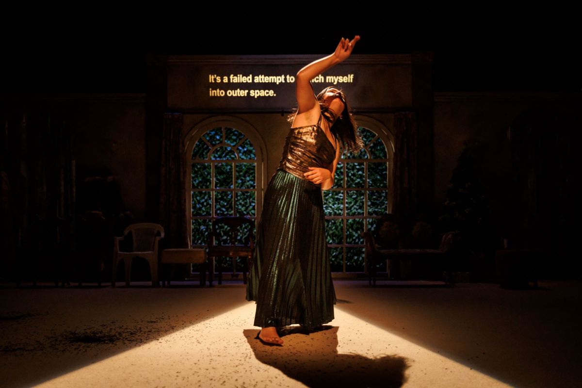 A person dances barefoot in a shaft of light. They wear a long green pleated skirt, a metallic bronze sleeveless top, and stand with one arm raised above their long hair and another hand on their hip.