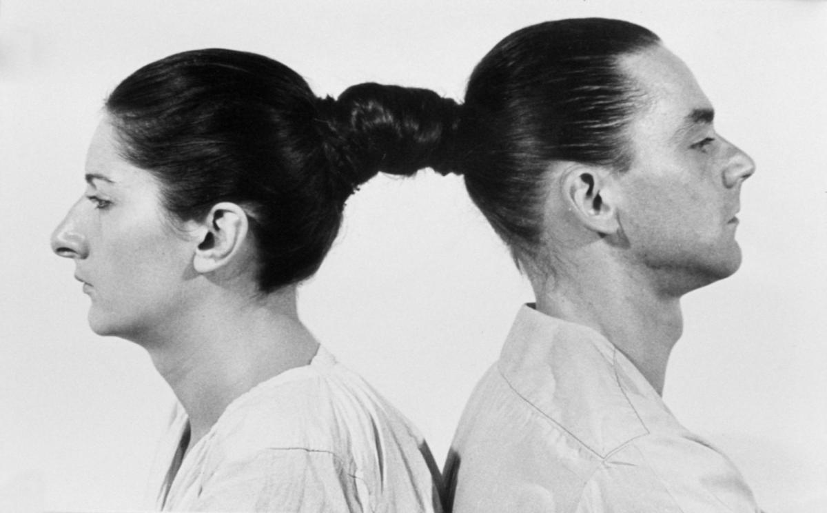 A black and white video still of a woman and a man back to back. They are connected by their hair, which is in a pony tail for both.