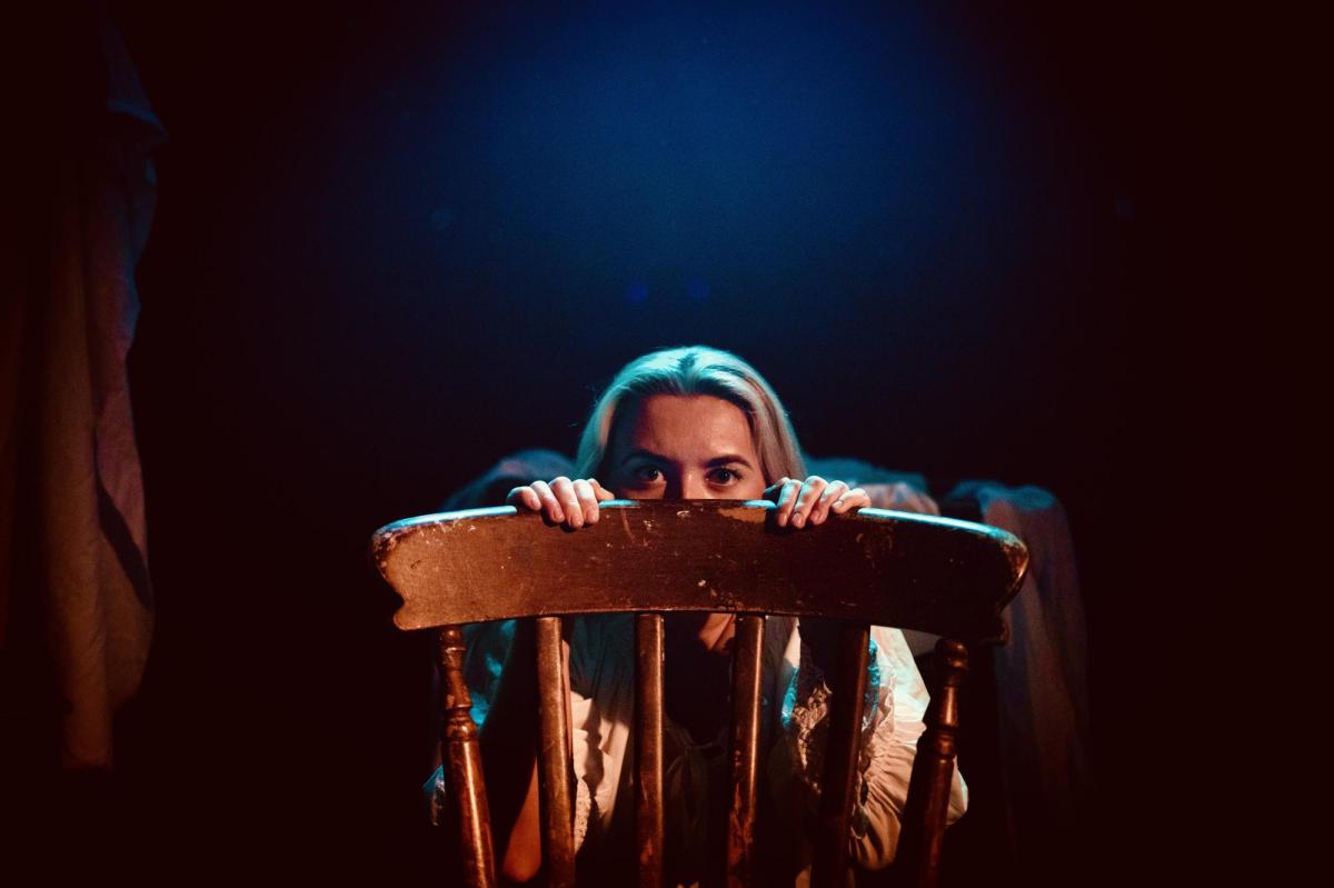 A blonde woman is peering over the top of a chair. You can only see her head and upper body.