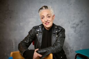 Ruth Mackenzie CBE leans on the back of a chair, smiling at the camera. She wears a black leather jacket over a black blouse, and her short white hair contains a few streaks of black.