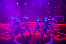 The six cast members of the musical 'Six' strut their stuff. They wear fluorescent-framed sunglasses and matching ruffs, and pose on a stage lit dramatically in red and purple.