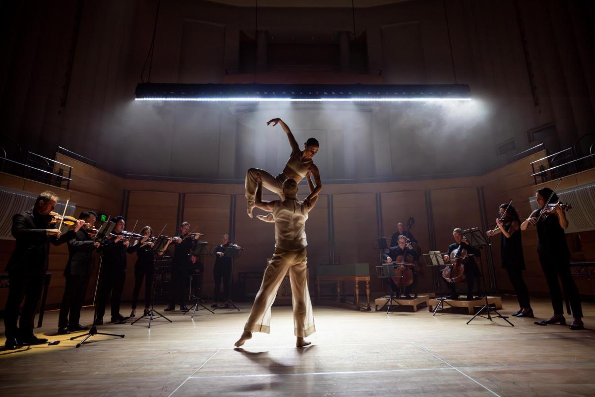 A couple in white are dancing. She is being lifted up in the air. Surrounding them is an orchestra of strings in black.