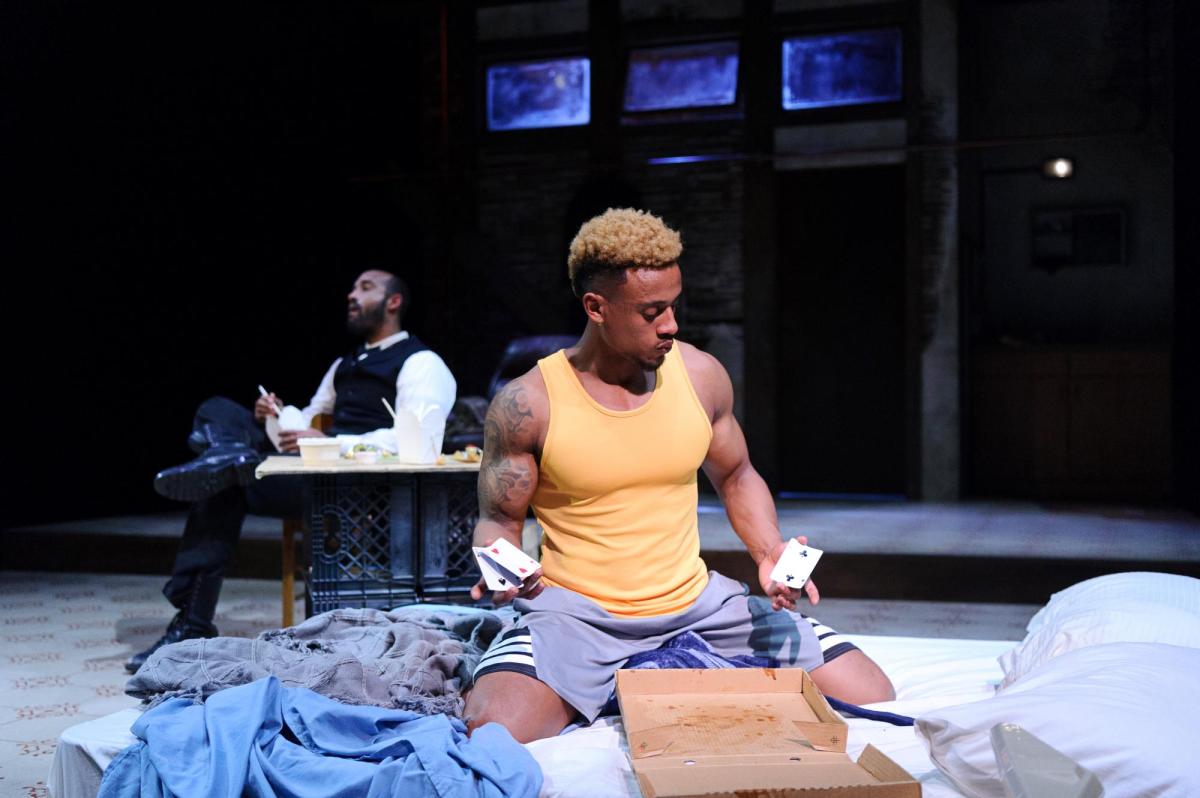 Two black men. Ras-Samuel is sitting on unmade bed with a pile of cards. He is wearing a yellow singlet and blue shorts. Damon Manns is in background seated, wearing a vest and white shirt. They are in the MTC play Top Dog/Underdog.