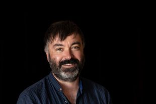 A dark-haired and bearded man in his 40s smiles at the camera. He wears a dark blue striped shirt and stands against a black background.